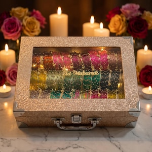 Puede incluir: Una caja decorativa dorada y brillante que contiene pulseras coloridas. La caja tiene una tapa transparente con la inscripción "Eid Mubarak" en letras doradas. Velas encendidas y flores en el fondo.