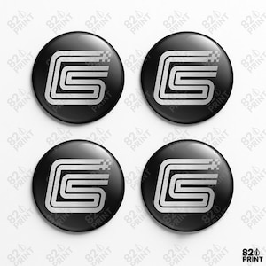 Puede incluir: Cuatro botones redondos negros con acabado brillante, cada uno con un diseño "S" blanco estilizado. El diseño incluye líneas paralelas y un elemento pixelado. Los botones están dispuestos sobre un fondo blanco.