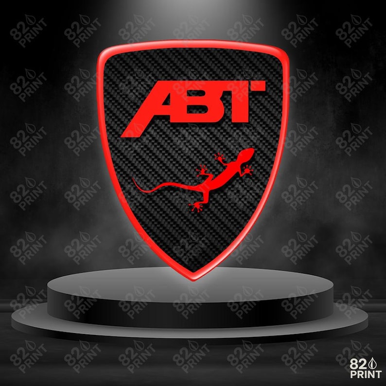 May include: A shield-shaped emblem with a carbon fiber background and a red border. The red text "ABT" is at the top, and a red lizard is at the bottom. The emblem is displayed on a gray platform.