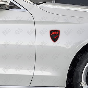 May include: A red and black ABT emblem affixed to the side of a white car. The emblem is shield-shaped with the ABT logo in red and a carbon fiber-like background. The car's body panel and tire are visible.