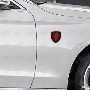 Pu&ograve; includere: Un emblema per auto nero e rosso a forma di scudo. L'emblema presenta un design a serpente rosso su uno sfondo nero in fibra di carbonio. L'emblema &egrave; attaccato al lato di un'auto bianca.