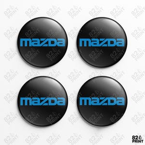 May include: Four black, circular Mazda emblems, each featuring the word "MAZDA" in blue. The emblems have a glossy finish and a domed shape. The background is white with a repeating pattern of "820 PRINT".