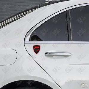May include: A red and black ABT emblem is affixed to the side of a white car. The emblem is shield-shaped with the ABT logo and a checkered pattern. The car's door handle and window are visible.