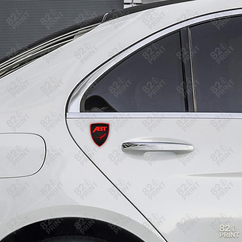 May include: A red and black ABT emblem is affixed to the side of a white car. The emblem is shield-shaped with the ABT logo and a checkered pattern. The car's door handle and window are visible.