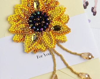 May include: A beaded sunflower brooch with yellow, gold, and dark blue beads. The brooch has dangling gold beaded strands with teardrop-shaped crystals. The words "For You" are printed on the white card.