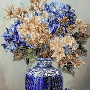 Puede incluir: Naturaleza muerta de cuentas con un jarrón azul oscuro lleno de un ramo de flores en tonos azules, crema y dorados. El jarrón tiene un borde de cuentas blancas detallado. Un ramo más pequeño está junto al jarrón, sobre un fondo neutro.