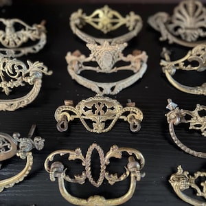 May include: A collection of antique brass drawer pulls with ornate designs. The pulls feature various shapes, including curved handles and decorative backplates. The brass has a tarnished, aged appearance, suggesting vintage or repurposed hardware.
