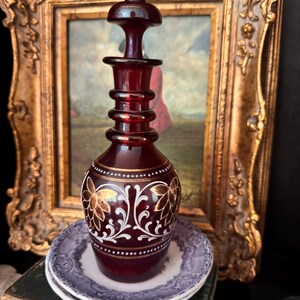 May include: A deep red glass decanter with a matching stopper, featuring a narrow neck with three horizontal rings and a bulbous base decorated with white scrollwork and gold floral motifs, resting on a small, light-colored ceramic saucer.