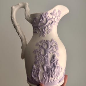 May include: A white ceramic pitcher featuring raised lavender-colored relief decorations. The handle is shaped like a tree branch. The body displays detailed scenes of figures on horseback and trees with acorns, finished with a matte texture.