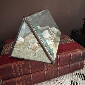 May include: A glass terrarium in a triangular shape, filled with sand and seashells. The terrarium sits atop two antique books with red covers. The books are resting on a white lace doily.