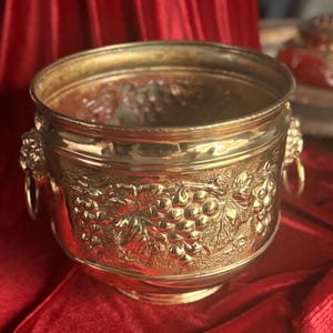 May include: A polished brass bucket featuring embossed grape clusters and vine leaf patterns on the exterior. The vessel includes two side handles shaped like lion heads with attached metal rings, providing a sturdy grip for this metallic container.