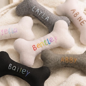Puede incluir: Una colección de seis juguetes de peluche para perros con forma de hueso en varios colores, incluyendo negro, gris y crema. Cada hueso presenta nombres bordados como Layla, Abne, Stella, Bentley, Bailey y Abby en hilos de distintos colores.