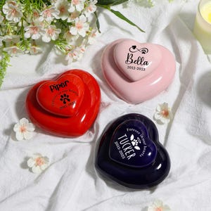 Op de afbeelding: Drie hartvormige urnen voor huisdieren in rood, roze en marineblauw. Elke urn heeft een kleiner hartje bovenop met de naam van een huisdier, data en een pootafdrukontwerp. De rode urn zegt "Piper".