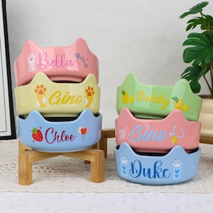 May include: A stack of six ceramic cat-shaped pet bowls in pastel colors. Each bowl is personalized with a pet's name in a playful font, along with decorative paw prints, fish bones, strawberries, and hearts. The bowls are displayed on a wooden stand.