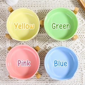 May include: Four colorful ceramic bowls, each with a different color and the color's name printed on it: yellow, green, pink, and blue. Each bowl has a small wooden handle on each side.