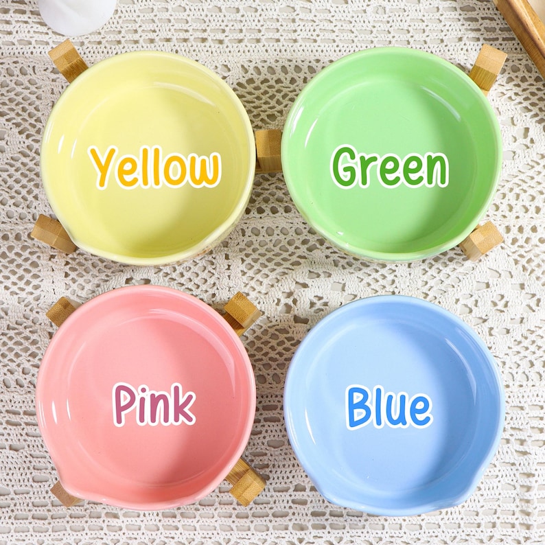 May include: Four colorful ceramic bowls, each with a different color and the color's name printed on it: yellow, green, pink, and blue. Each bowl has a small wooden handle on each side.