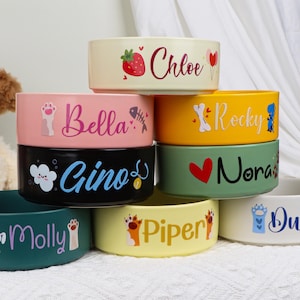 Puede incluir: Una pila de cuencos de cerámica para mascotas en varios colores, cada uno con el nombre de una mascota en escritura. Los cuencos presentan elementos decorativos como huellas de patas, corazones y gráficos relacionados con la comida. Los colores incluyen crema, rosa, amarillo, negro, verde y turquesa.