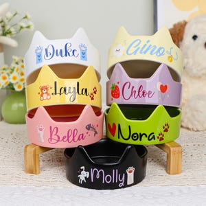 Puede incluir: Una pila de seis cuencos de cerámica para mascotas en varios colores, cada uno con un diseño de oreja de gato. Los cuencos están personalizados con nombres como "Duke" y "Molly", junto con huellas de patas decorativas y otras pequeñas ilustraciones. Los cuencos se exhiben en un soporte de madera.