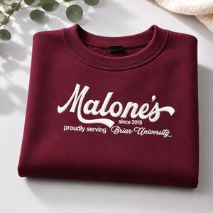 May include: A maroon crewneck sweatshirt featuring white embroidered text that reads Malone's since 2015 proudly serving Briar University. The garment has a ribbed collar and a soft, fleece-lined interior visible at the neckline.