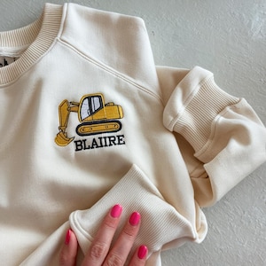 May include: Cream-colored sweatshirt featuring a yellow embroidered excavator design with the name "BLAIRE" in black. The sweatshirt has ribbed cuffs and a crew neck. The image shows a close-up of the garment.