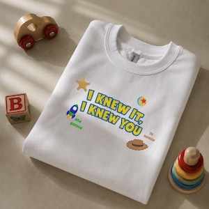 May include: A white crewneck sweatshirt featuring embroidered text that reads I KNEW IT, I KNEW YOU, along with small colorful icons including a yellow star, a blue and green rocket, a red and yellow ball, and a brown cowboy hat with text.