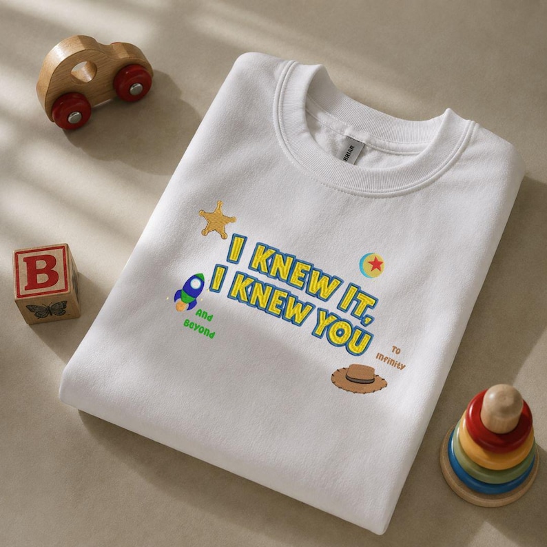 May include: A white crewneck sweatshirt featuring embroidered text that reads I KNEW IT, I KNEW YOU, along with small colorful icons including a yellow star, a blue and green rocket, a red and yellow ball, and a brown cowboy hat with text.