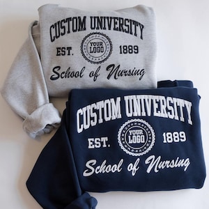 Peut inclure: Deux sweat-shirts, un gris et un bleu marine, avec le texte "CUSTOM UNIVERSITY" et "School of Nursing". Chaque sweat-shirt porte un logo circulaire et l'année "1889". Le sweat-shirt gris est au-dessus du bleu marine.