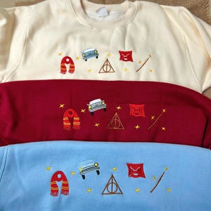 May include: Three sweatshirts in cream, red, and light blue, each embroidered with a scarf, car, triangle, envelope, and wand. The embroidery is in gold and black. The sweatshirts are folded on a neutral surface.
