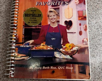 May include: A spiral-bound cookbook titled "MY FAMILY'S FAVORITES" featuring a woman in a red top and blue apron. The cover includes a "NOW A BESTSELLER" sticker and the text "OVER 175,000 IN PRINT". The author is Mary Beth Roe, QVC Host.