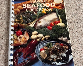 May include: A spiral-bound cookbook titled "Hawaii Seafood Cookbook" with a blue cover featuring images of seafood dishes, fish, and vegetables. The author's name, Howard Hansen, is printed at the bottom.