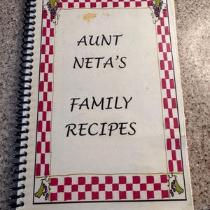 以下が含まれることがあります： 「Aunt Neta's Family Recipes」と題された、スパイラル綴じの料理本。表紙は白地で、赤と白のチェック柄の縁取りが施されています。タイトルは黒い文字で、隅にはブドウの装飾画が描かれています。