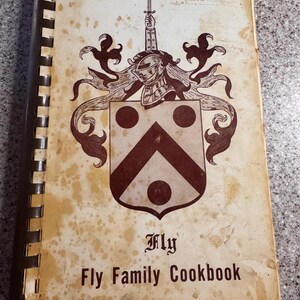 Puede incluir: Libro de cocina vintage titulado "Fly Family Cookbook". La portada presenta un escudo familiar en marrón y blanco, con un yelmo y una espada. El libro tiene encuadernación en espiral y muestra signos de edad con manchas.