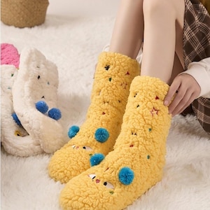 Könnte beinhalten: Gelbe, flauschige Socken mit Cartoon-Gesichtern, blauen Pompons und bunten Sternen. Die Socken werden an den Beinen einer Person getragen, wobei sich im Hintergrund weitere weiße und rosa Socken befinden. Die Socken sind aus einem weichen, plüschigen Material.