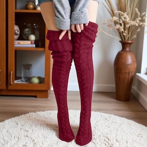 May include: Burgundy cable knit over-the-knee socks. These thick, textured socks have a folded cuff at the top. The socks, in a deep red hue, are designed to be worn over the knees. The cable knit pattern adds a classic touch to the design.