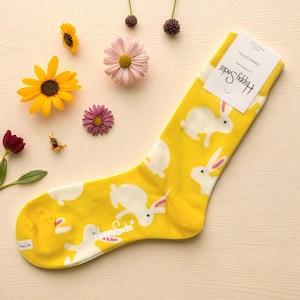 May include: A yellow sock with a white bunny pattern. The sock has the words "Happy Socks" printed on it. The sock is surrounded by flowers and a small bee.