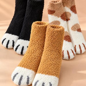 Puede incluir: Tres pares de calcetines tobilleros de felpa con diseños de patas de animales. Un par es negro sólido, otro es marrón claro sólido y otro es blanco con manchas marrones. Cada calcetín tiene una punta blanca con detalles de almohadillas.