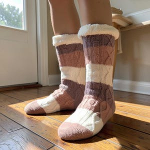 May include: A pair of mid-calf knit socks featuring a cable knit pattern in alternating horizontal stripes of dusty rose, mauve, and cream. The socks are finished with a thick, white faux-fur cuff at the top opening, providing a textured look.