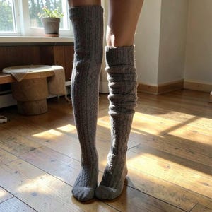 May include: A pair of over-the-knee socks in a heathered grey knit fabric. The socks feature a vertical cable knit pattern running the length of the leg. One sock is pulled straight up, while the other is bunched slightly around the calf area.