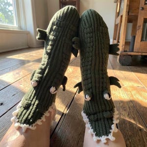 Peut inclure: Une paire de chaussettes en tricot vert olive foncé en forme de crocodiles. Les chaussettes présentent un motif côtelé texturé rappelant des écailles, de petites nageoires saillantes sur les côtés, des dents crochetées blanches et des yeux.