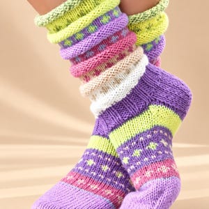 May include: A pair of mid-calf knit socks featuring a multi-coloured striped pattern in shades of lime green, purple, pink, and white. The socks have a thick, textured ribbed cuff and a repeating geometric floral motif throughout the leg and foot.