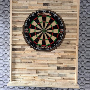 Op de afbeelding: Een houten dartbordkast met een Winmau dartbord. De kast is gemaakt van verschillende stukken hout, gerangschikt in een mozaïkpatroon. Het dartbord heeft zwarte, rode, groene en witte kleuren. Onderaan de kast bevindt zich een klein plankje.