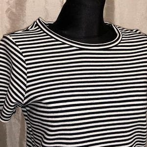 May include: A black and white striped t-shirt with a crew neck and short sleeves. The shirt is displayed on a black mannequin head. The stripes are horizontal and evenly spaced, creating a classic nautical look.