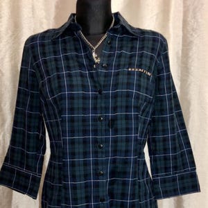 May include: A dark green and navy blue plaid button-down shirt with three-quarter sleeves. The shirt has a collared neckline and a small pocket detail. A silver necklace with a pendant is visible.