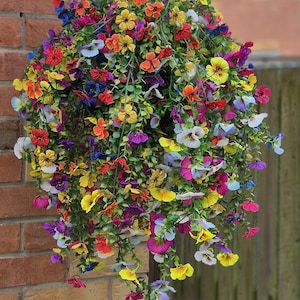 Peut inclure: Un panier suspendu débordant de fleurs artificielles colorées. Les fleurs sont un mélange de rouge, jaune, orange, bleu et violet, créant un affichage vibrant. Le panier est suspendu à un support en métal noir sur un mur de briques.
