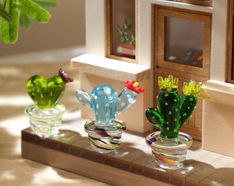 Puede incluir: Tres figuras de cactus de vidrio en macetas decorativas. Los cactus son verdes, azules y amarillos con detalles rojos y amarillos. Las macetas tienen diseños a rayas coloridas. Las figuras se exhiben sobre una superficie de madera frente a un edificio en miniatura.