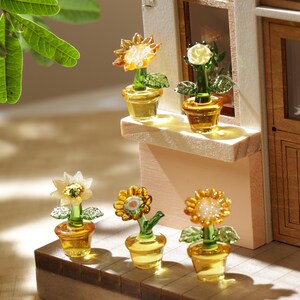 Może przedstawiać: Miniaturowe szklane kompozycje kwiatowe w kolorze żółtym i zielonym. Kwiaty, w tym słoneczniki i róże, znajdują się w małych żółtych doniczkach. Scena rozgrywa się na drewnianej powierzchni, z widokiem na miniaturowy budynek w tle.