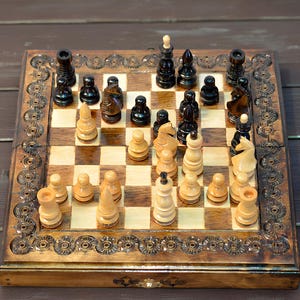 Könnte beinhalten: Ein Schachspiel aus Holz mit detailliertem, geschnitztem Rand. Die Schachfiguren sind schwarz und hellbraun, angeordnet auf einem karierten Brett. Das Set wird auf einer dunklen Holzoberfläche präsentiert.