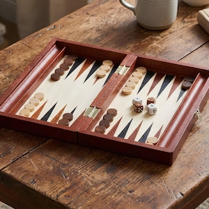 May include: A wooden backgammon set with a dark brown frame and a light-colored playing surface. The set includes brown and beige game pieces, dice, and is open on a wooden table. The game is ready to play.
