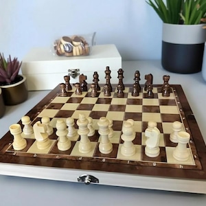 Puede incluir: Un juego de ajedrez de madera con casillas marrones claras y oscuras y piezas de ajedrez. Las piezas de ajedrez están dispuestas en el tablero, listas para jugar. Una caja blanca y una bolsa de damas están en el fondo.