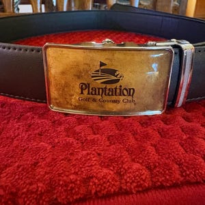 Puede incluir: Un cinturón de cuero marrón oscuro con una hebilla rectangular plateada y una inserción brillante de color tostado. La inserción muestra un gráfico negro de una bandera de golf y el texto Plantation Golf & Country Club.